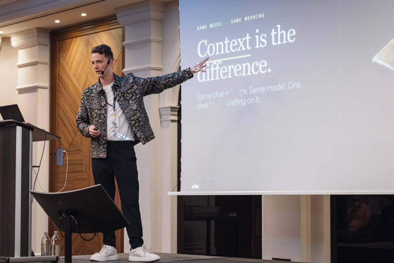 Speaker on stage, audience in low light — a webinar recording.